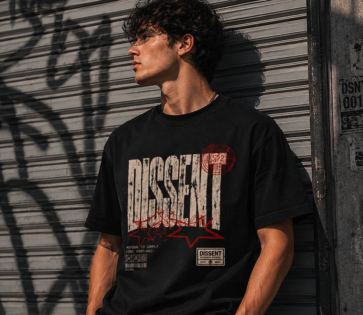 A man wearing a black graphic t-shirt with a distressed white and red 'DISSENT' logo, leaning against a gray metal shutter door with graffiti.